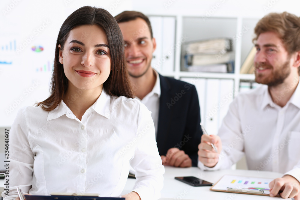 Beautiful smiling cheerful girl at workplace look in camera with colleagues group in background. White collar worker at workspace job offer modern lifestyle client visit profession train concept