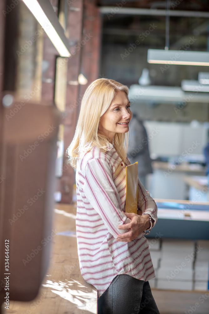 Fototapeta premium Side view of blonde woman standing at the window, holding yellow folder