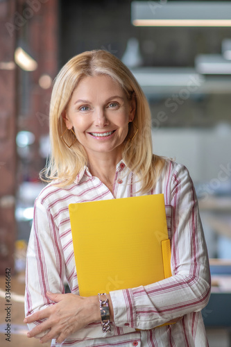 Blonde woman standing, holding yellow folder, smiling photo