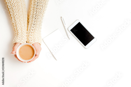Top view a young girl with her right hand holding a coffee Cup on a white table next to a phone with an empty area on the white cover in the morning