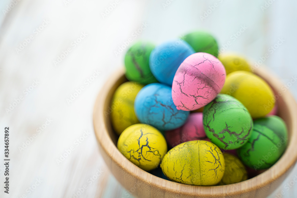 close up colorful vibrant easter egg of pink green yellow and blue colors piling in brown wooden bowl, on wooden table with white scraped paint background, representing celebration of easter holidays close up colorful vibrant easter egg of pink green yellow and blue colors piling in brown wooden bowl, on wooden table with white scraped paint background, representing celebration of easter holidays