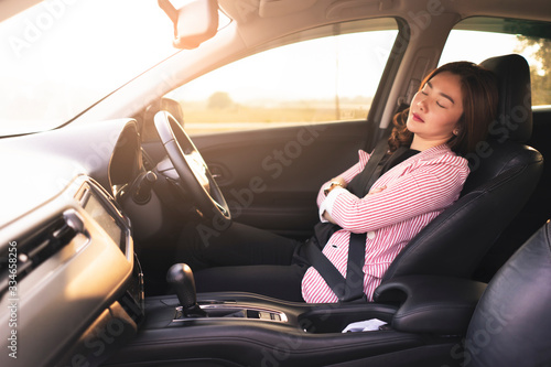 asian businesswoman laying sleeping and with the drivers set, with a seatbelt on resting in car with arms across the chest, black leather interior in the car and sunset shining through the car window © Have a nice day 