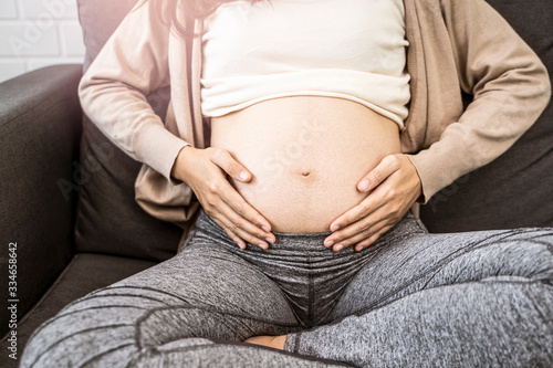 Close up asian pregnant woman placing hands on baby lump stomach feeling baby heartbeat, sitting on sofa relaxing and resting from tiredness, in living room with brick texture wall and white curtains