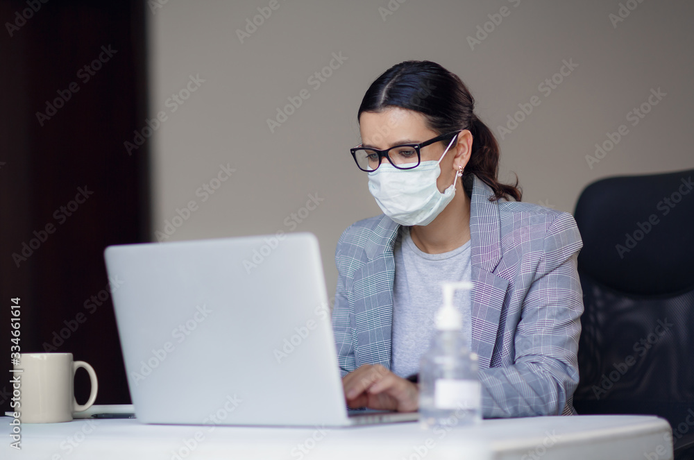Young female employee in medical protective mask from corona virus covid-19 working on laptop at workplace. Pandemic concept, contagious disease, virus outbreak.