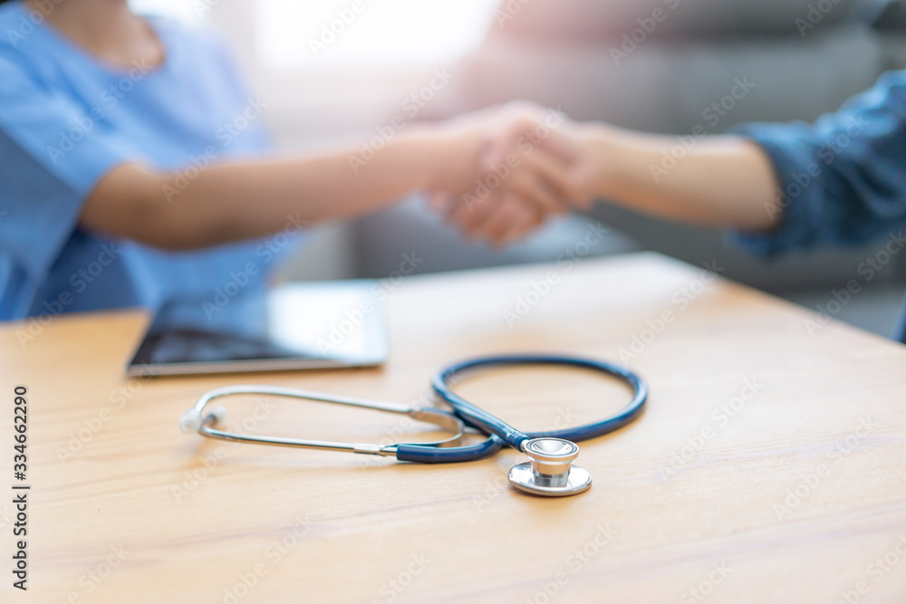 close up showing a stethoscope on table with tablet device and background of female doctor shaking the patients hand in greeting, goodbye and thankful for doctor looking after the patient in comfort close up showing a stethoscope on table with tablet device and background of female doctor shaking the patients hand in greeting, goodbye and thankful for doctor looking after the patient in comfort