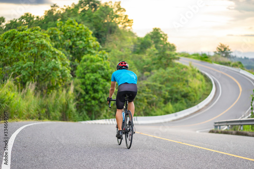 back view of a cyclist on top of a mountains winding road, riding a black bicycle down a hill, wearing bike helmet and blue cycling jersey, with grey clouds sunset sky and forest in the background. © Have a nice day 