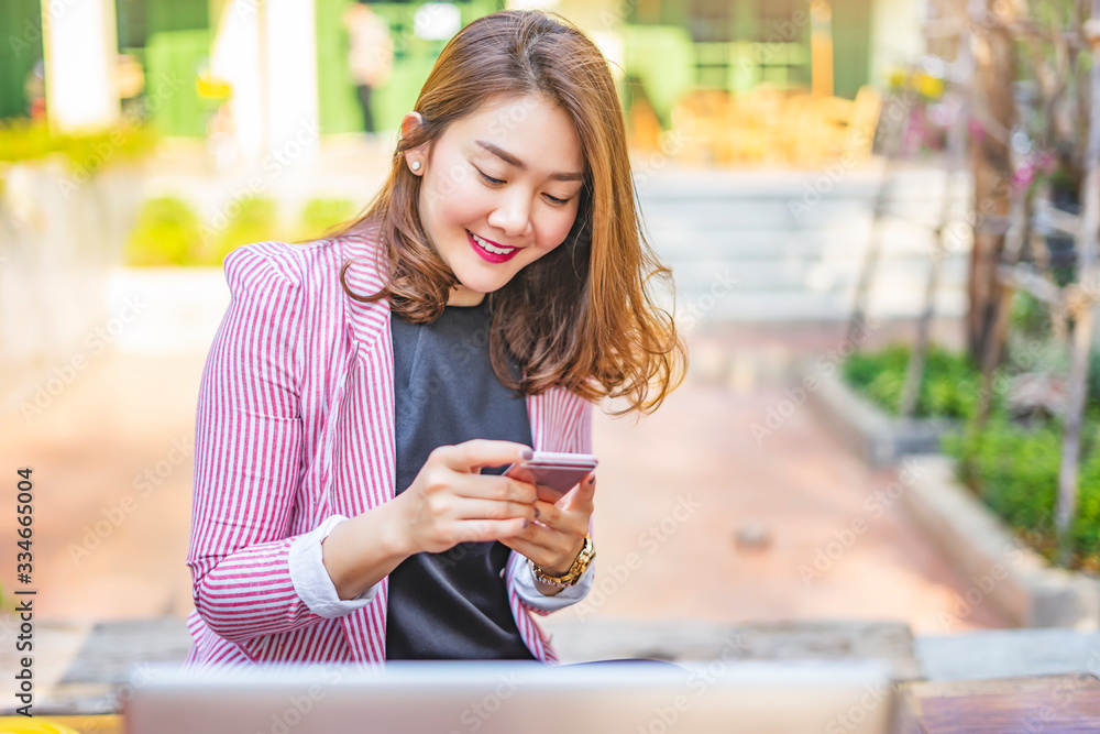 portrait of asian business woman holding and playing on her smartphone, wearing a watch, a pink and white stripe blazer, and black shirt, smiling and sitting one a wooden table in the summer sunshine portrait of asian business woman holding and playing on her smartphone, wearing a watch, a pink and white stripe blazer, and black shirt, smiling and sitting one a wooden table in the summer sunshine