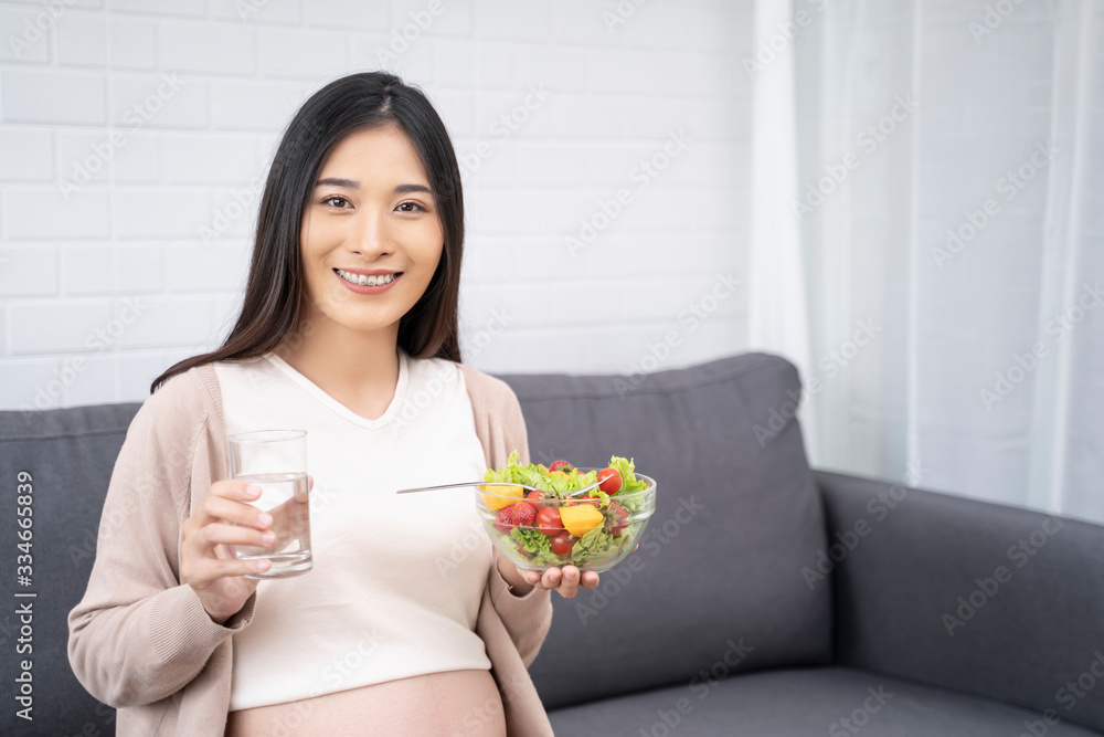 Beautiful asian pregnant woman holding a bowl of salad of vegetables and fruits, using a fork eating tomato. Eating healthy having lunch sitting on the grey sofa, relaxing and resting living room Beautiful asian pregnant woman holding a bowl of salad of vegetables and fruits, using a fork eating tomato. Eating healthy having lunch sitting on the grey sofa, relaxing and resting living room