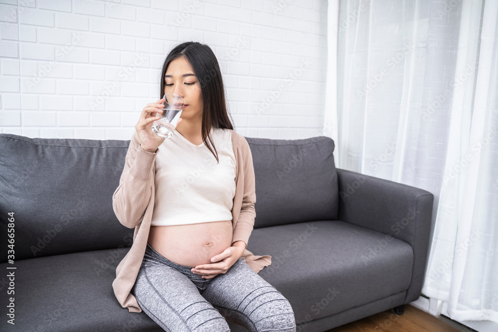 Beautiful asian pregnant woman drinking holding glass of water, eating healthy having lunch sitting on the grey sofa, smiling joyfully relaxing and resting living room with white brick wall texture Beautiful asian pregnant woman drinking holding glass of water, eating healthy having lunch sitting on the grey sofa, smiling joyfully relaxing and resting living room with white brick wall texture