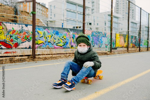 boy in a medical mask, jacket and khaki hat sits on an orange longboard on the street against a backdrop of graffiti and a metal fence