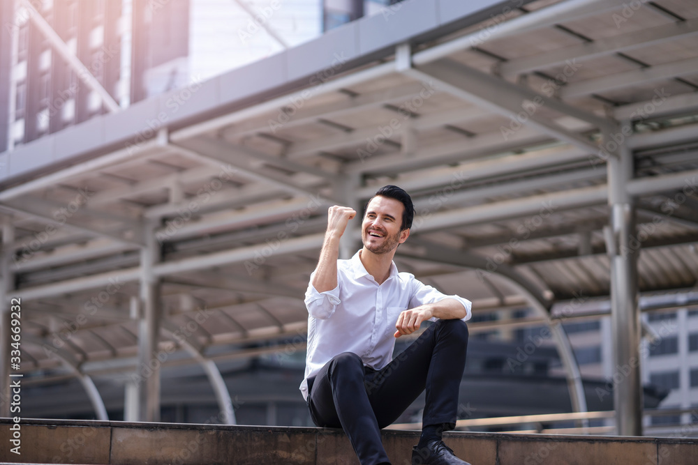 male business worker sitting on top of stairway with fist thrusted up and smiley face feeling happy, representing success or happiness in a workplace, in the city with modern architectural structure.. male business worker sitting on top of stairway with fist thrusted up and smiley face feeling happy, representing success or happiness in a workplace, in the city with modern architectural structure..