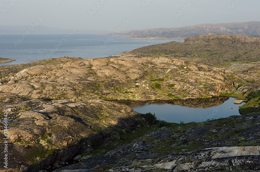 Sunny warm summer arctic landscape with sea view and blue lake - slope of granite shore and calm Barents Sea.