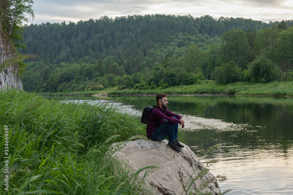Tourist man enjoy with beautiful view of nature of forest, river, mountains. Young bearded guy sitting on big stone on mountain river bank