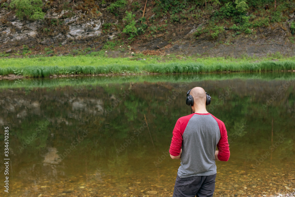 Rear view of bald young man standing on river bank and listening to music in headphones. Far from big city bustle. Relaxing outdoors. Melomaniac