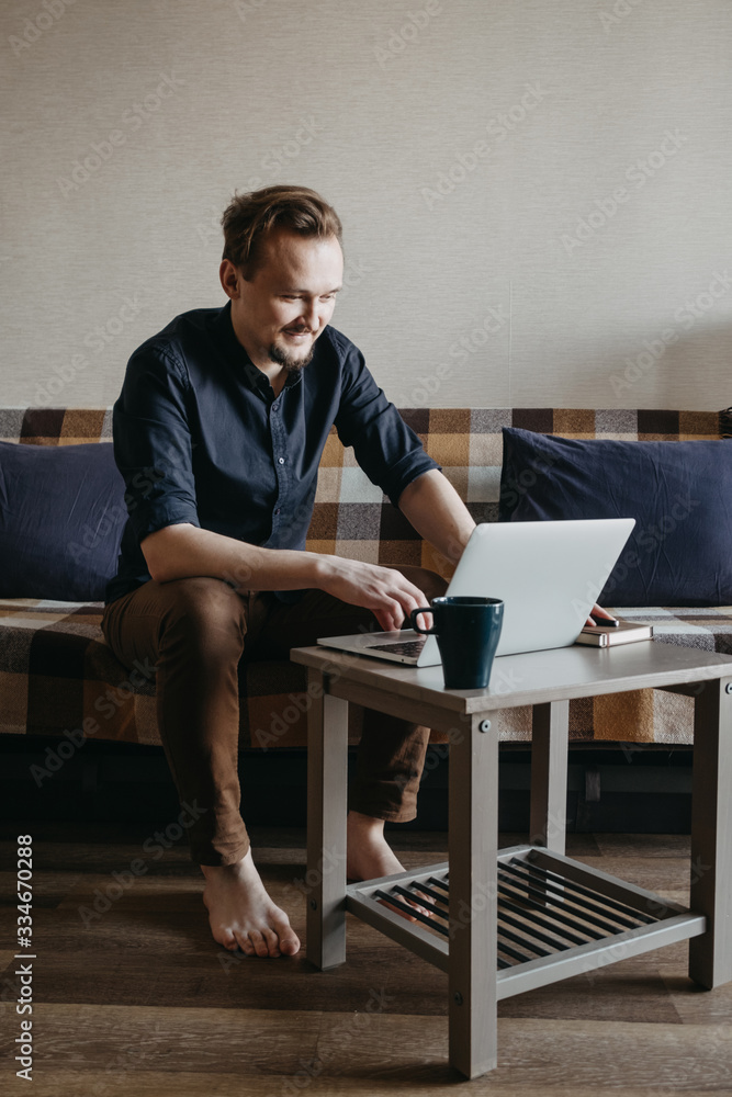 A young European guy in a dark blue shirt and brown trousers with a dark beard looks at a laptop. Sits on a sofa, laptop a small table. Home insulation. Home Office.