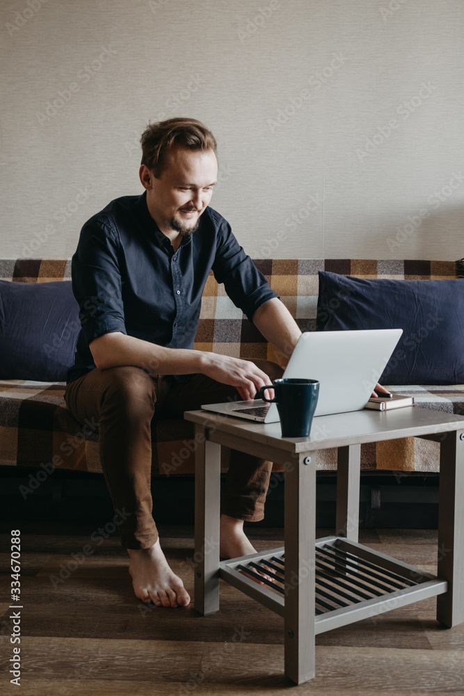 A young European guy in a dark blue shirt and brown trousers with a dark beard looks at a laptop. Sits on a sofa, laptop a small table. Home insulation. Home Office. A young European guy in a dark blue shirt and brown trousers with a dark beard looks at a laptop. Sits on a sofa, laptop a small table. Home insulation. Home Office.