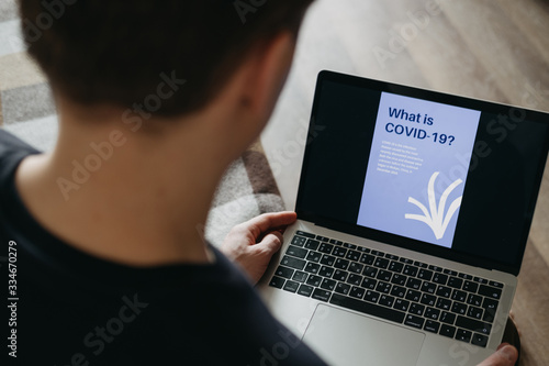 A young guy looks at the laptop screen, where it says about coronovirus. Photo taken over the shoulder of a young man