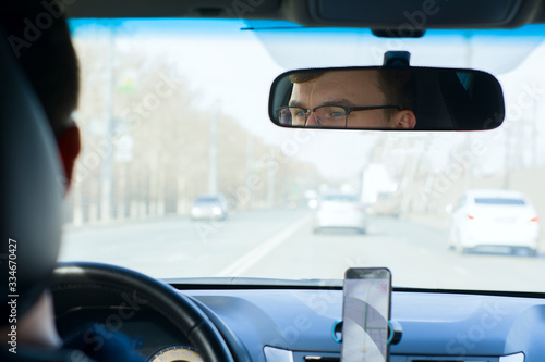 a young man with glasses at the wheel of a car is reflected in the rearview mirror