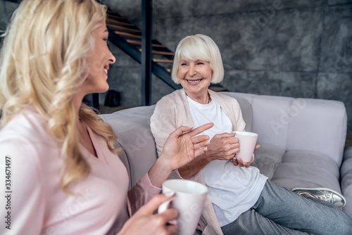 Senior woman and her attractive daughter spending time together at home. Sitting on sofa and drinking tea together. Happy Mothers' Day.