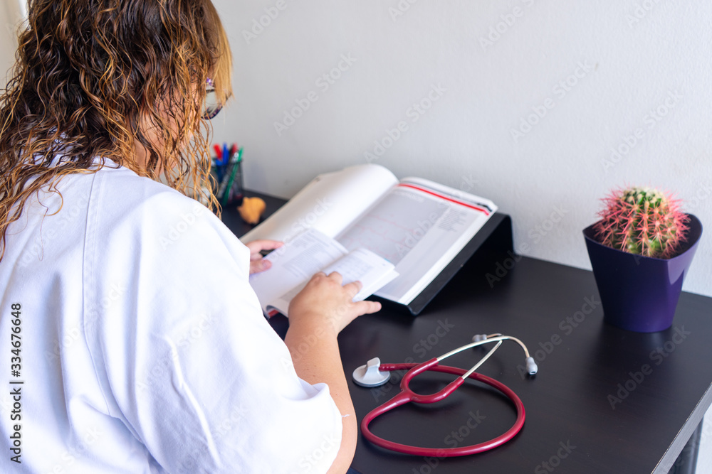 Young woman studying medicine sitting at a desk. There is also a cardiology textbook where you can see electrocardiograms. On the desk there is a stethoscope and a cactus. Young woman studying medicine sitting at a desk. There is also a cardiology textbook where you can see electrocardiograms. On the desk there is a stethoscope and a cactus.