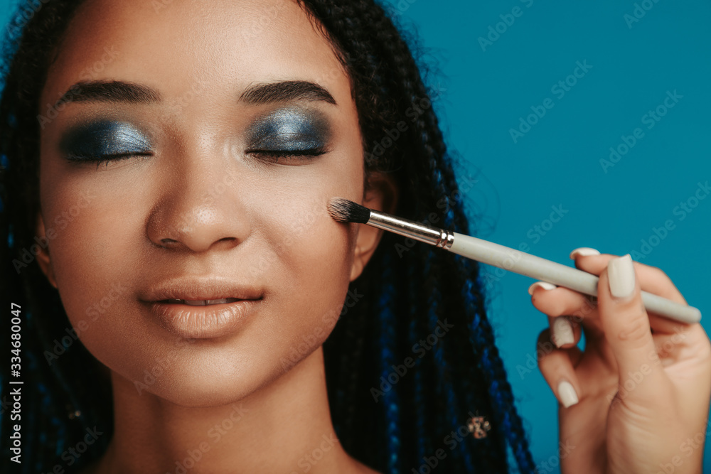 Obraz premium Calm peaceful attractive afro american woman with closed eyes on photo. Hand hold make up brush and touch model's cheek. Eyeshadows with blue metal color. Isolated over blue background.