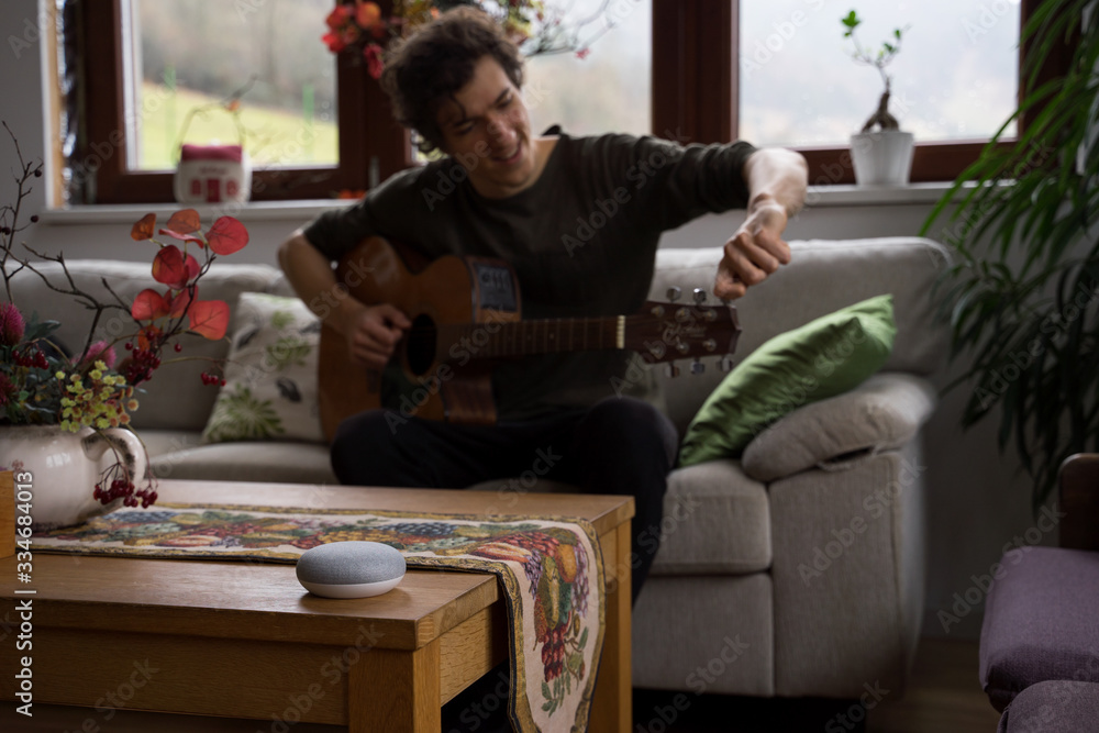 smart home voice activated speaker in home with man on guitar in background. Stay at home.