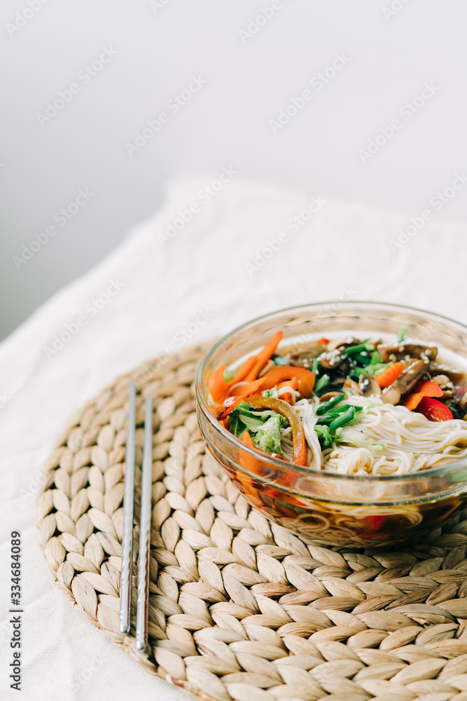 Japanese ramen soup in a glass plate with vegan ingredients, fresh vegetables in small plates, rice noodles, mushrooms and miso paste broth, on a natural wicker napkin on a white table Japanese ramen soup in a glass plate with vegan ingredients, fresh vegetables in small plates, rice noodles, mushrooms and miso paste broth, on a natural wicker napkin on a white table