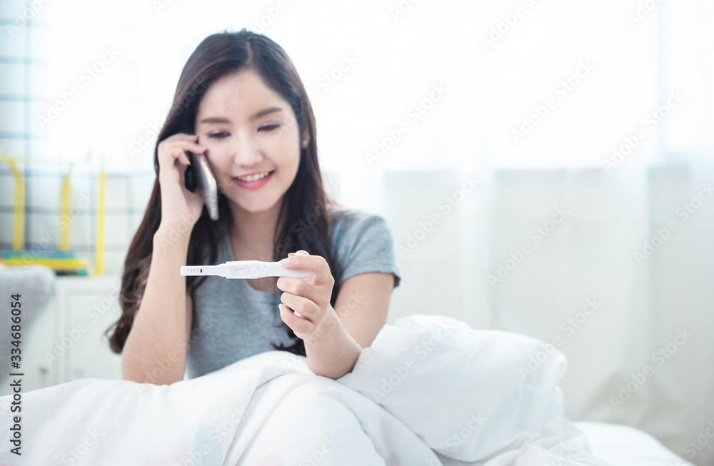 Young beautiful asian woman using smart mobile phone hand holding positive pregnancy test with smile and happy sitting on bed in bedroom at home. Young beautiful asian woman using smart mobile phone hand holding positive pregnancy test with smile and happy sitting on bed in bedroom at home.