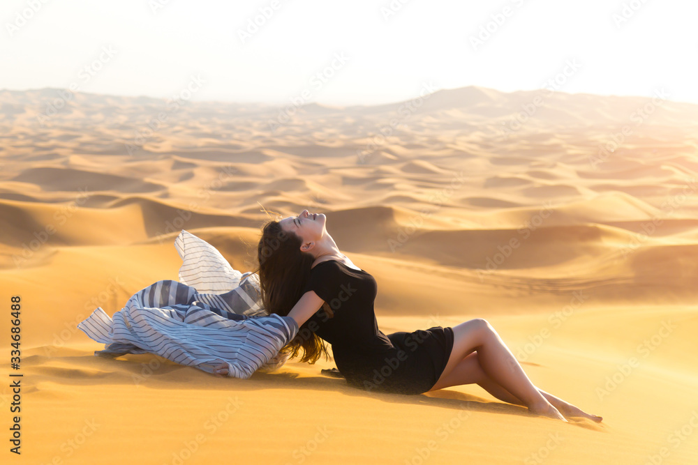 Young model in black dress posing lying on the sand in the desert. Beautiful young woman gracefully posing in the sand.