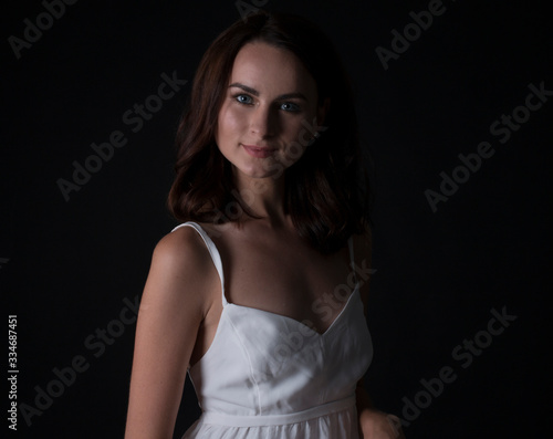 stunning portrait of a sexy young brunette girl in a white dress sitting on a high chair on a black background. Low key. Beauty and Fashion