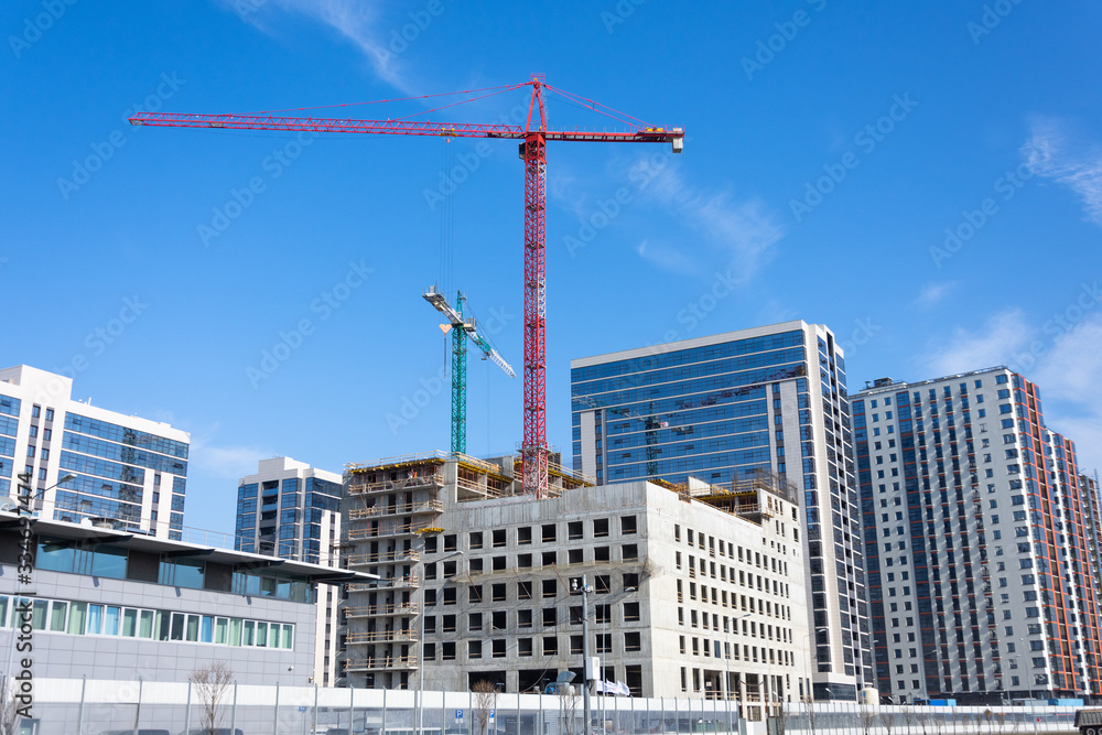 View of a building with high construction cranes and nearby a glass tower, built houses. View of a building with high construction cranes and nearby a glass tower, built houses.