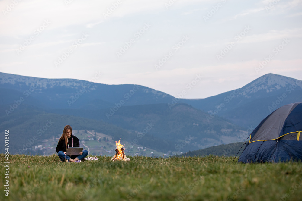 young female freelancer working on laptop in the mountains in the evening. Tourist girl sitting near campfire and having fun. Copy space.