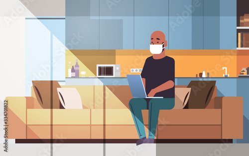 african american man in protective mask using laptop coronavirus pandemic quarantine concept work from home online training education freelance modern living room interior full length horizontal