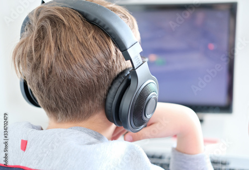 teenager boy looking at computer monitor. happy smiling boy in earphones playing computer games. over shoulder view.Distance learning concept.