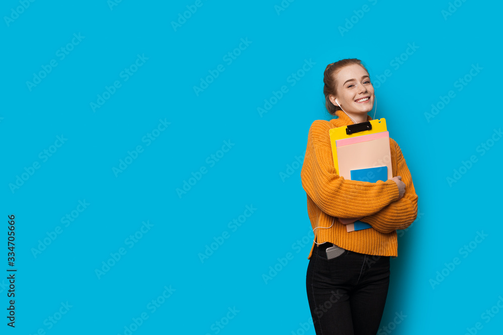 Lovely caucasian student with red hair and freckles is posing with earphones and some books on a blue wall with free space Lovely caucasian student with red hair and freckles is posing with earphones and some books on a blue wall with free space