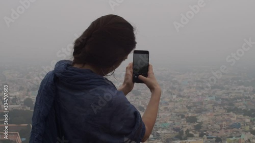 Wallpaper Mural Back view of young woman making photo of misty cityscape on telephone outdoors copy text space. Unrecognizable brunette girl holding mobile phone shooting panoramic city slow motion. Social media Torontodigital.ca