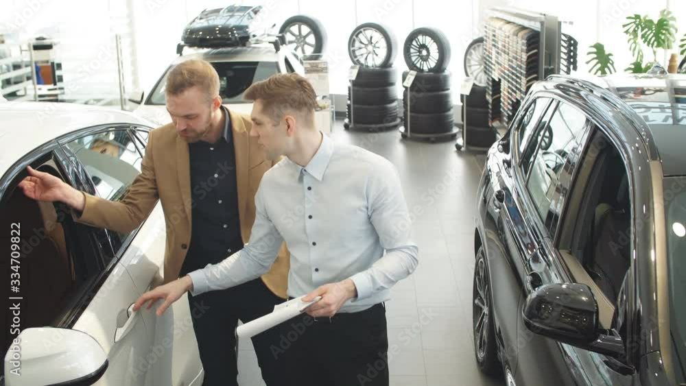 Young affable consultant talking about features of new car to customer. Business man listen to him, choosing new auto in dealership. Young affable consultant talking about features of new car to customer. Business man listen to him, choosing new auto in dealership.