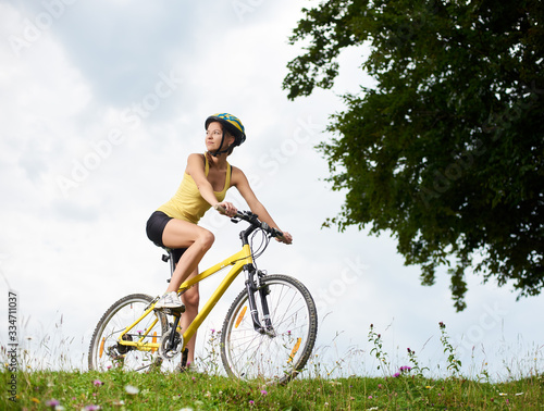 Attractive happy woman cyclist riding on yellow mountain bicycle on a grassy hill, wearing helmet, enjoying summer day in the mountains. Outdoor sport activity, lifestyle concept