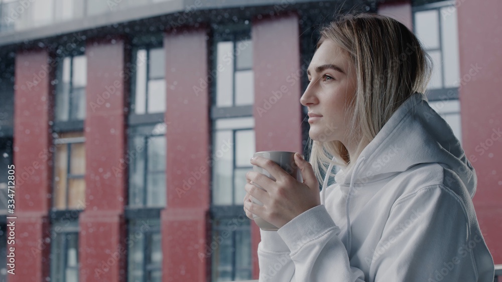 Pretty european young woman in white hoodie staying in balcony with cup of hot coffee or tea relax enjoy breathing fresh air healthy freezy snow falls in march morning no stress concept Pretty european young woman in white hoodie staying in balcony with cup of hot coffee or tea relax enjoy breathing fresh air healthy freezy snow falls in march morning no stress concept