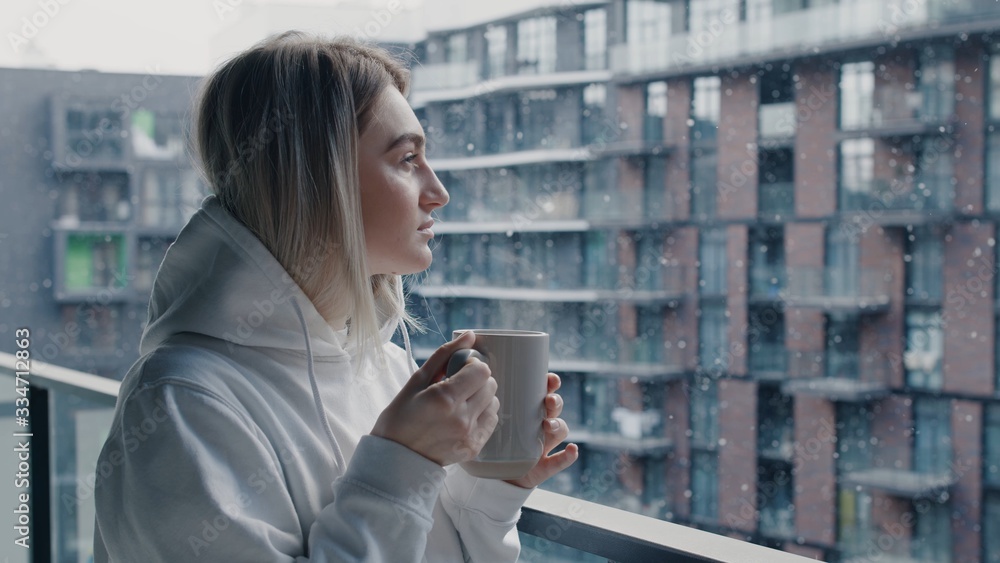 Close up view of cheerful young woman in white hoodie stays in balcony drink hot coffee or tea relax enjoy breathing fresh air healthy freezy snow falls in march morning no stress concept Close up view of cheerful young woman in white hoodie stays in balcony drink hot coffee or tea relax enjoy breathing fresh air healthy freezy snow falls in march morning no stress concept