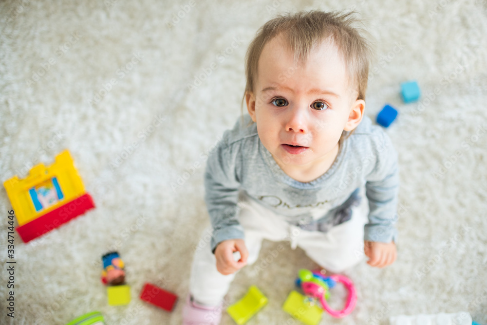 Portrait of one year old baby girl indoors in bright room siting on a carpet.