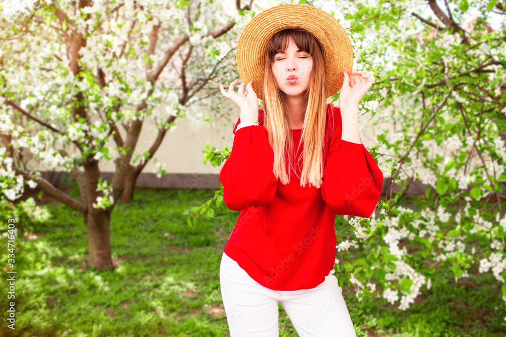 Pretty blonde asian woman in straw hat, white jeans and red boho shirt posing over pink blooming tree in sunny spring day. Holidays and shopping concept.