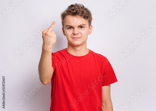 Portrait of angry teen boy showing middle finger, on grey background. Handsome caucasian young teenager showing bad gesture. Upset cute child doing obscene sign.