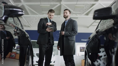 Manager in auto dealership presents new cars using a tablet. Standing in a car dealership customer is listening to manager.