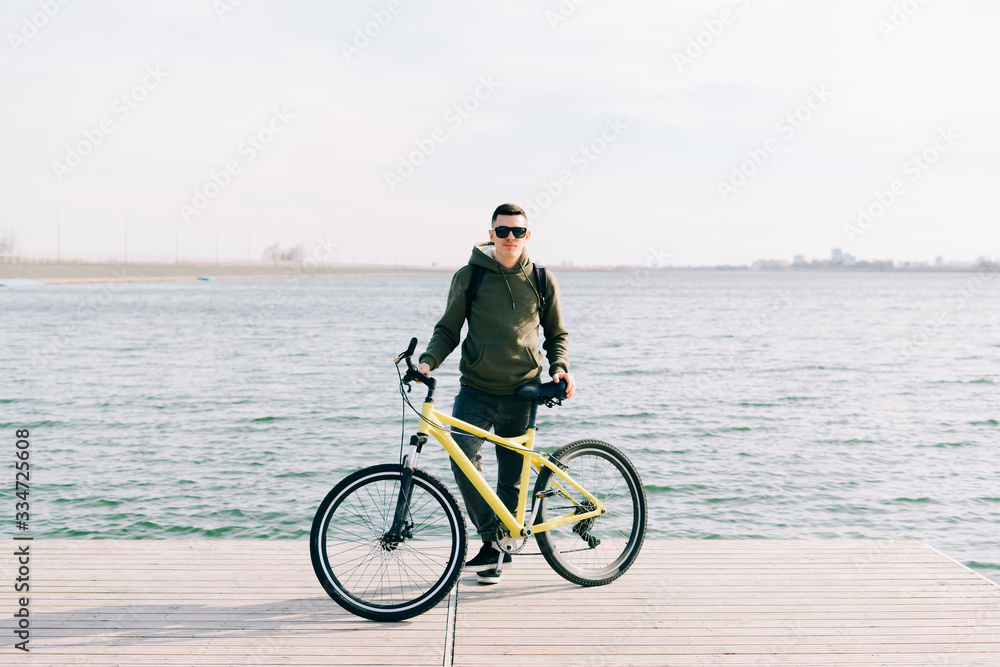 a young man in khaki sportswear and glasses stands on a pontoon near the lake and holds a yellow Bicycle near him on a Sunny spring day a young man in khaki sportswear and glasses stands on a pontoon near the lake and holds a yellow Bicycle near him on a Sunny spring day