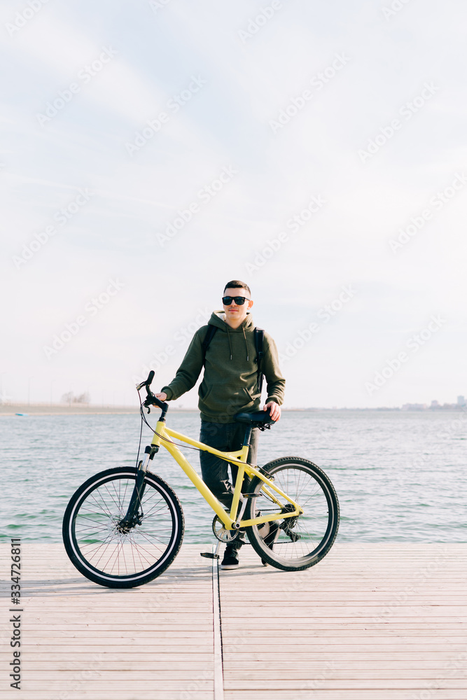 a young man in khaki sportswear and glasses stands on a pontoon near the lake and holds a yellow Bicycle near him on a Sunny spring day