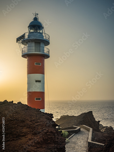 Weg aus Steinen zum rot wei  en Leuchtturm Punta de Teno auf Teneriffa im Gegenlicht bei Sonnenuntergang mit Meer und Dunst