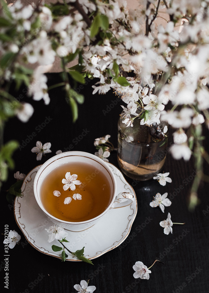 On a black background is a cup of tea next to a vase filled with flowering branches On a black background is a cup of tea next to a vase filled with flowering branches