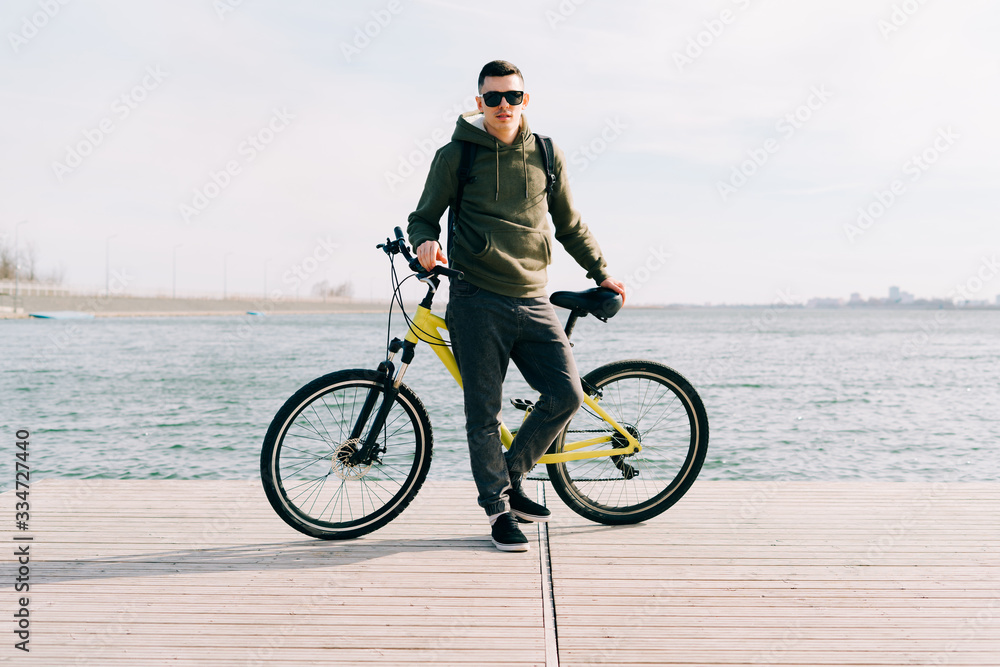 Obraz premium a young man in khaki sportswear and glasses stands on a pontoon near the lake and holds a yellow Bicycle near him on a Sunny spring day