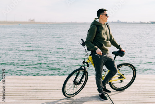 a young man in khaki sportswear and glasses stands on a pontoon near the lake and holds a yellow Bicycle near him on a Sunny spring day